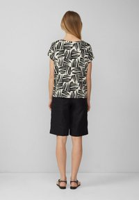 Korte mouwen top in roomkleur met zwart bladerenpatroon, gecombineerd met zwarte linnen shorts. Sandalen zijn plat met dunne banden. Achteraanzicht weergegeven.
