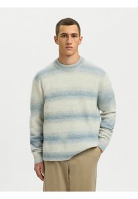Jeune homme portant un pull en tricot à rayures bleu clair et blanc et un pantalon beige, debout devant un fond blanc uni.