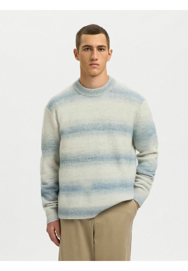 Jeune homme portant un pull en tricot à rayures bleu clair et blanc et un pantalon beige, debout devant un fond blanc uni.