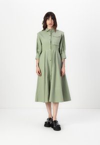 Rochie verde deschis, cu nasturi, fustă plisată, guler de cămașă și mâneci trei sferturi. Prezintă un buzunar pe piept și un material neted.