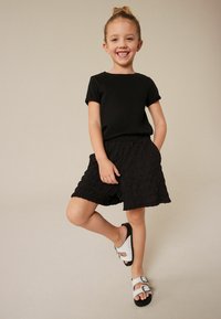 Camiseta negra, pantalones cortos negros texturizados y sandalias blancas con hebillas. Niña posando con una pierna doblada, manos en los bolsillos de los pantalones cortos. Fondo sencillo.