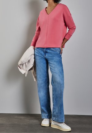 Personne portant un haut rose à col en V, un jean bleu clair, des baskets blanches, tenant une veste beige sur un fond neutre.