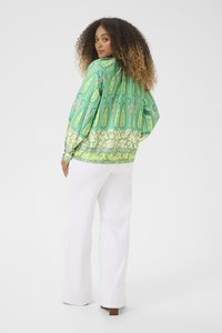 Blusa verde con motivo paisley e maniche lunghe, caratterizzata da disegni intricati. Abbinata a pantaloni bianchi a gamba larga e scarpe con tacco marroni.