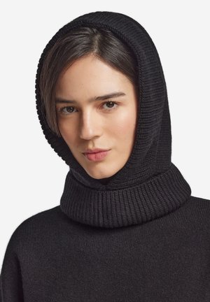 Ung kvinde med mørkt hår iført en sort ribstrikket hættetrøje, der vender front mod kameraet med et neutralt udtryk mod en hvid baggrund.