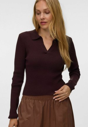 Femme aux longs cheveux blonds portant un haut à manches longues côtelé marron foncé avec un col et une jupe marron sur un fond uni.