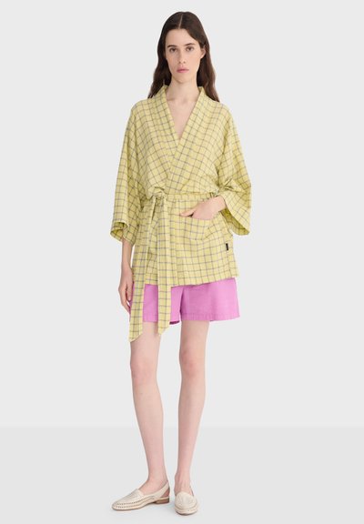 Femme portant un haut cache-cœur jaune à carreaux avec de larges manches, un short rose et des chaussures crème sans lacets, debout devant un fond clair uni.