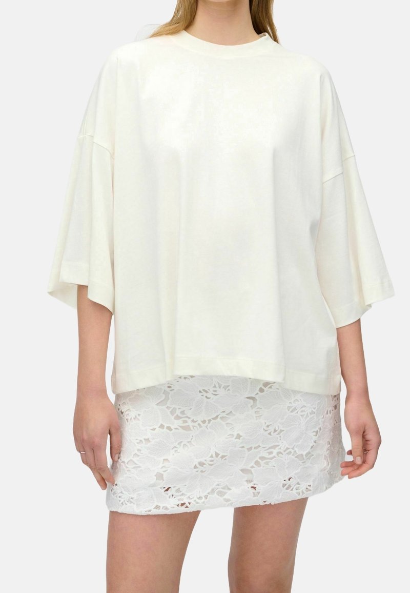 Femme portant un T-shirt blanc ample et oversize et une jupe courte en dentelle blanche, debout sur un fond uni et clair.
