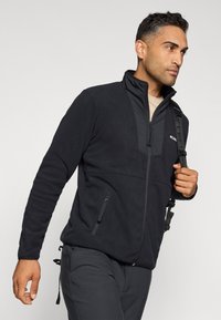 Black fleece jacket with a high collar, zippered front, and two side pockets. Features a textured material and contrast stitching.