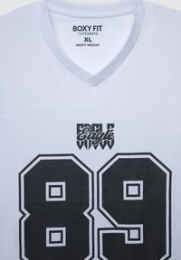 Biała, gruba koszulka z dekoltem w serek, z wyrazistymi czarnymi grafikami: "Orzeł" oraz numerem "89" obrysowanym na biało. Prosty, luźny krój.