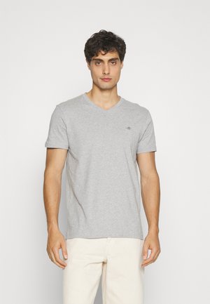 Mann trägt ein schlichtes hellgraues V-Ausschnitt-T-Shirt und elfenbeinfarbene Hose, steht vor einem einfarbigen weißen Hintergrund.