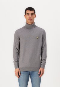Grijze coltrui gemaakt van zachte stof, met een klein geel logo op de borst en ribbelstructuur bij de manchetten en de onderkant.