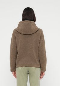 Καφέ φλις μπουφάν με ψηλό γι ακά και υπερμεγέθη κουκούλα, με υφή από απαλό, μαλακό υλικό και χαλαρή εφαρμογή, φορεμένο πάνω από ανοιχτό πράσινο παντελόνι.