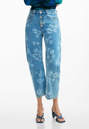 Vaqueros azules recortados de talle alto con estampado floral claro, cierre de botones y combinados con sandalias azules de tacón sobre pies descalzos.