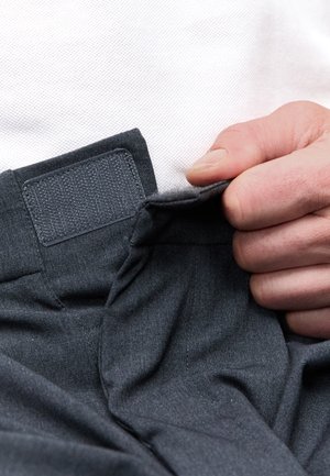 Hand houdt en opent zwarte stoffen tailleband met klittenbandsluiting, wit overhemd op de achtergrond.