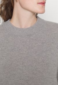 Frau trägt einen grauen Rundhalsstrickpullover und goldene Creolen, schaut nach rechts mit sichtbarem Kiefer und Lippen.