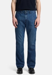 Uomo che indossa jeans in denim blu a gamba dritta, cintura nera, scarpe nere e una maglia scura in piedi contro uno sfondo bianco semplice.