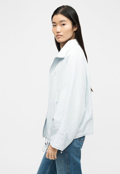 Νεαρή γυναίκα με μακριά σκούρα μαλλιά που φοράει ανοιχτό μπλε σακάκι με φερμουάρ και μπλε τζιν, στραμμένη ελαφρώς πλαγίως σε λευκό απλό φόντο.