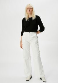 Zwarte geribde trui, witte cargo-broek met wijde pijpen en zijzakken, en crèmekleurige enkelboots. Glad materiaal met een relaxed fit en knoopsluiting.