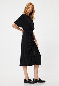 Zwarte jurk met een ontspannen top, gatherend taille en uitsnijding. Glad materiaal, kniehoogte, gecombineerd met zwarte instappers. Minimalistisch ontwerp.
