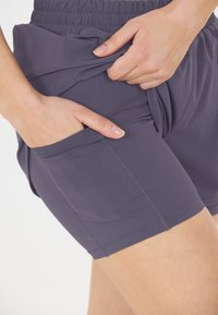 Lila sportshorts med elastisk midja, med en sidoficka och jämn textur. Stygnsynliga sömmar längs kanterna.
