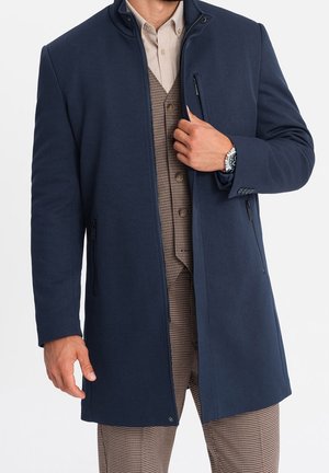 Muž v tmavě modrém dlouhém kabátě přes béžovou košili a hnědý kostkovaný oblek, upravuje přední část kabátu u kapsy se zipem.