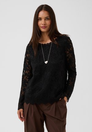 Mujer con el pelo oscuro y largo que lleva una blusa negra de encaje de manga larga, pantalones marrones y un collar con colgante plateado, de pie contra un fondo liso.