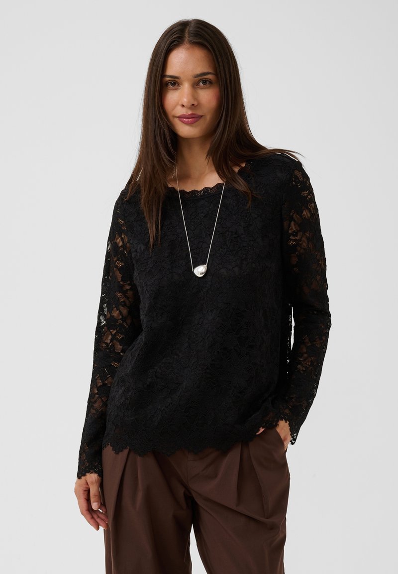 Femme aux longs cheveux foncés portant un haut en dentelle noire à manches longues, un pantalon marron et un collier avec un pendentif argenté, debout devant un fond uni.