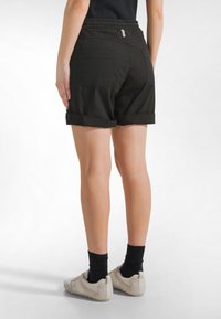 Frau trägt schwarze hochgekrempelte Shorts, schwarze Socken, beige Sneaker, steht mit einer Hand auf der Hüfte, Rücken zur Kamera vor weißem Hintergrund.