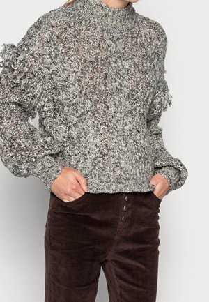 Fleckiger, strukturierter Strickpullover in Grautönen mit hohem Kragen und Fransenverzierungen an den Ärmeln, kombiniert mit braunen Cordhosen mit Knöpfen.
