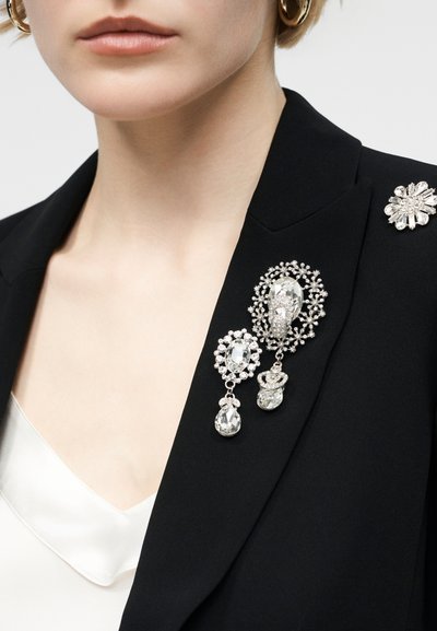 Γυναίκα που φοράει μαύρο σακάκι με τρία περίτεχνα ασημένια κρυστάλλινα καρφίτσες και λευκό σατέν τοπ από κάτω, εμφανιζόμενη από τον λαιμό έως το στήθος.