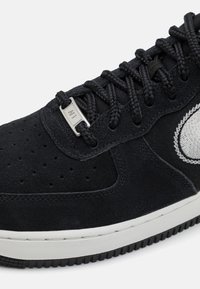 Adidași din suede negru cu suport metalic pentru șireturi, vârf perforat, accente texturate și talpă din cauciuc alb. Detaliu cu logo sportiv în gri.