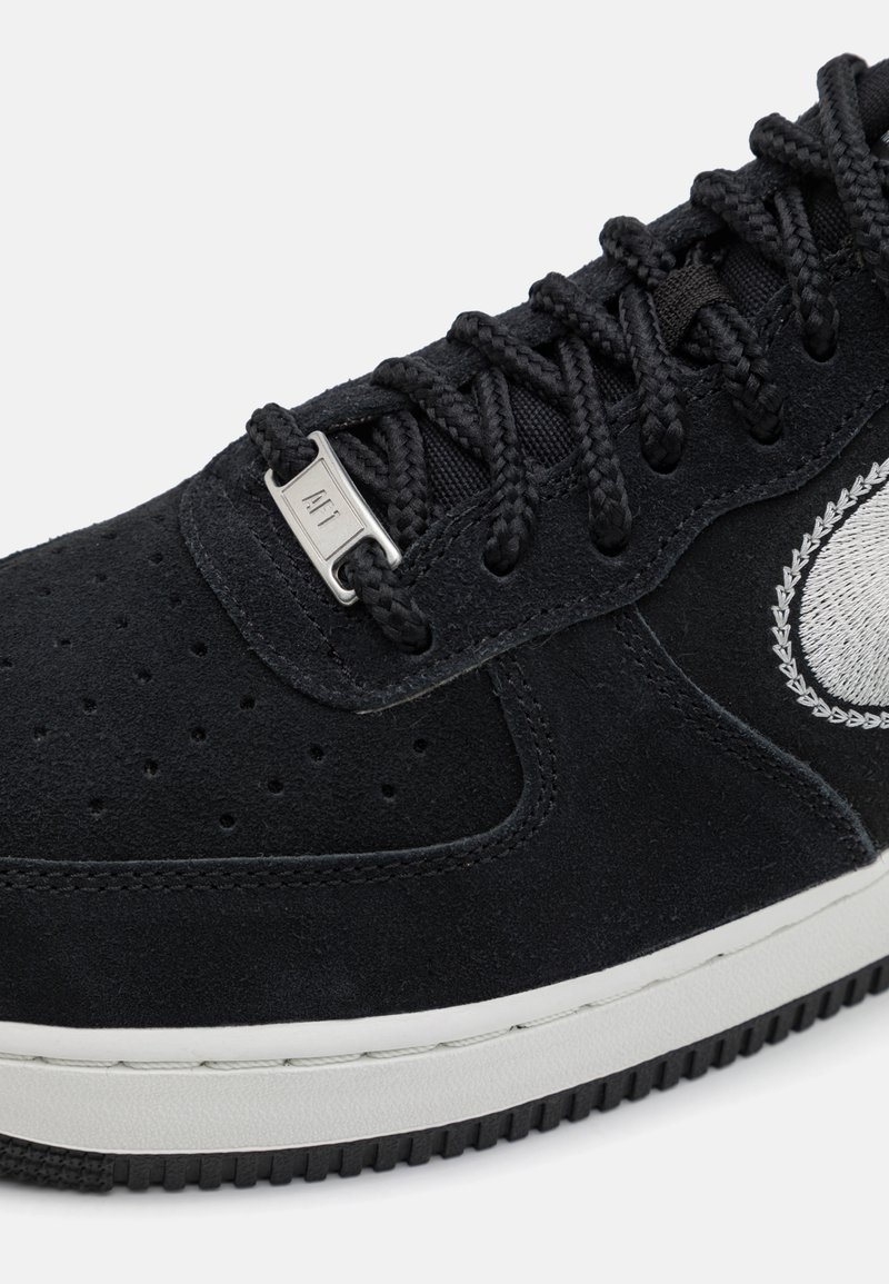 Zwarte suède sneakers met metalen vetersluiting, geperforeerde neuzen, textuuraccenten en een witte rubberen zool. Sportlogo-detail in grijs.