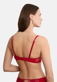 Frau mit langen braunen Haaren, die einen roten Spitzen-BH und passende Unterwäsche trägt, von hinten vor einem einfachen weißen Hintergrund gezeigt.