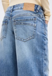 Jasnoniebieskie dżinsy z denimu z tylnymi kieszeniami i brązową skórzaną metką z napisem „perfect jeans gina tricot”, noszone z beżową bluzką z długim rękawem.