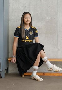 Jovem mulher sentada num banco de madeira contra uma parede de betão, vestindo uma camisola desportiva preta, saia plissada, meias brancas e ténis.