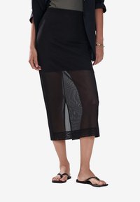 Gonna nera lunghezza al ginocchio con un soprabito trasparente a motivo, abbinata a sandali neri piatti. Tessuto liscio e leggero.