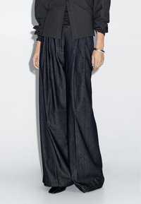 Dark denim wide-leg trousers with a high waist, featuring pleats for added volume and smooth texture, paired with a black striped top.