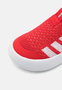 Røde sneakers med et pustende strikket overdel, hvite teksturerte striper og en glatt gummisåle. Har en polstret krage og hæltab.