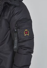 Schwarze Kapuzenjacke mit gepolstertem Textur, Reißverschlusstasche am Ärmel und gesticktem Emblem mit goldenen Akzenten. Logoetikett an der Tasche angebracht.