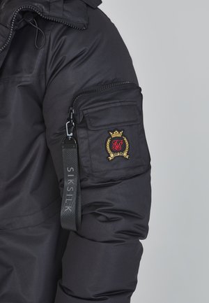 Black insulated jacket with a zippered sleeve pocket, featuring a gold and red embroidered logo and a strap labeled "SIK SILK."