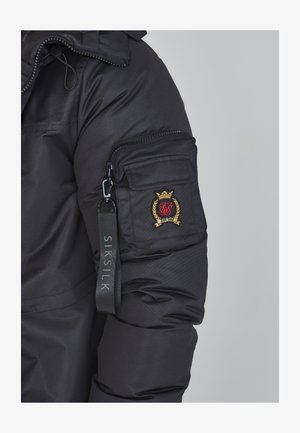 Must jakt, millel on lukuga käepide ja mis sisaldab kuldset ja punast broderiyeeritud logo ning riba, millel on märge "SIK SILK."