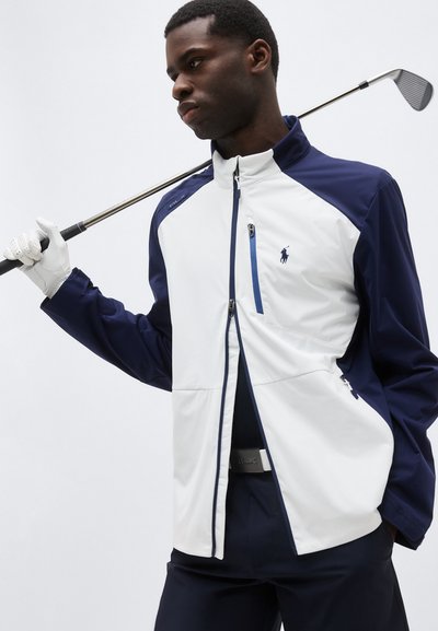 Hombre con chaqueta de golf azul marino y blanca y guante, sosteniendo un palo de golf sobre el hombro, mirando hacia un lado contra un fondo liso.