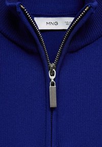 Niebieski sweter na zamek z wysokim kołnierzem, wyposażony w srebrny zamek błyskawiczny i mały metalowy suwak do otwierania i zamykania.