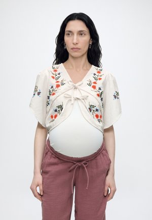 Gravid kvinde med langt mørkt hår iført en hvid blomsterbroderet cropped top over en tætsiddende hvid skjorte og løstsiddende mauve bukser.