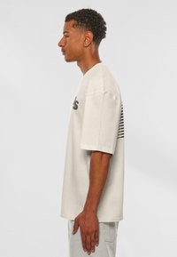 Cream-colored oversized T-shirt with a round neckline, short sleeves, and black horizontal stripes on the back, made from soft cotton fabric.