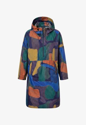 Femme portant une veste oversize colorée à motif abstrait, un jean bleu à revers et des bottines bleu foncé, debout les mains dans les poches.