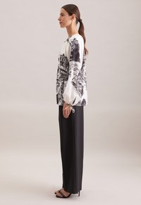 Donna in piedi di lato, che indossa una camicia bianca con stampe architettoniche nere, pantaloni larghi neri e sandali con tacco neri.