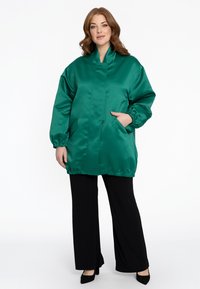 Groene satijnen jas met een staande kraag, elastische mouwen, zijzakken en een losse fit, gecombineerd met zwarte wijde broek en hakken.
