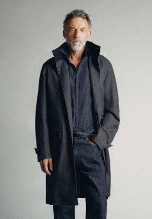 Man with gray beard wearing a dark overcoat and buttoned shirt, standing with one hand in pocket against plain background.