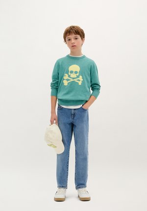 Türkiser Pullover mit gelbem Totenkopf und gekreuzten Knochen-Design, blaue Jeans, weiße Mütze in der Hand gehalten und weiße Sneaker mit braunen Sohlen.